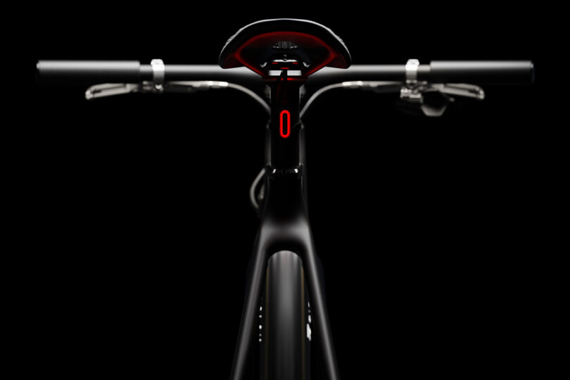 Close-up of a black bicycle viewed from the front, featuring handlebars, seat, and a red digital display on the frame against a black background.